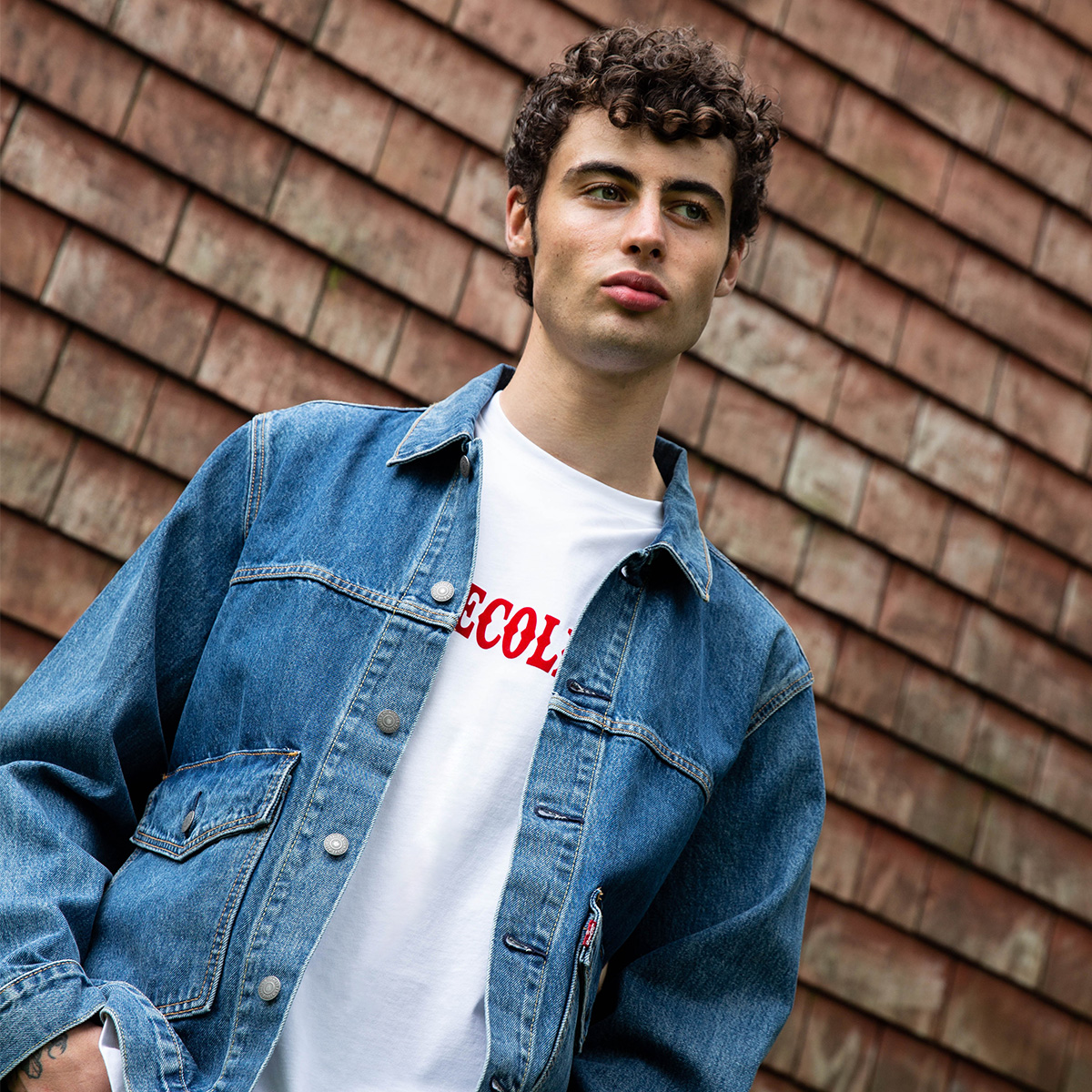 A man stood infront of a tiled wall. Wearing a Levis denim jacket and a white t-shirt from Versace Collection