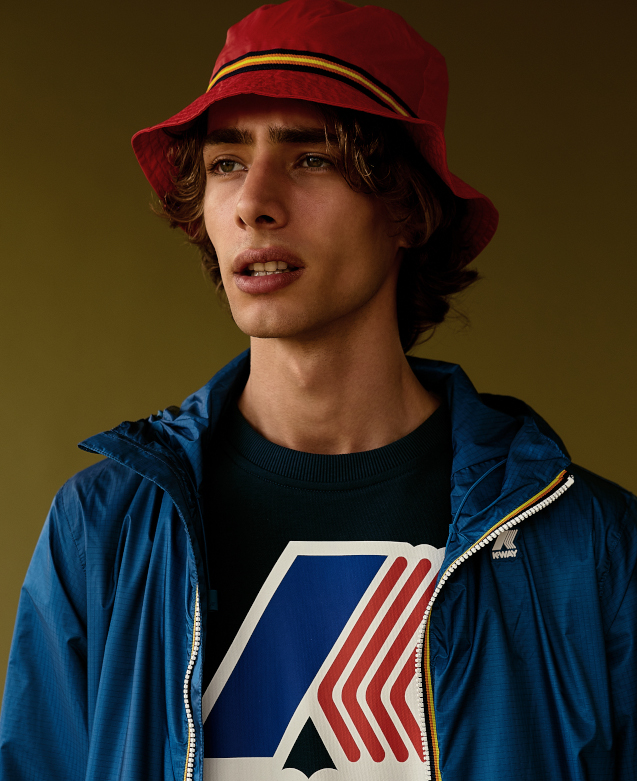 A young man is looking beyond the camera. He is wearing a blue k-way rain jacket, a red bucket hat and a black K-Way logo tshirt