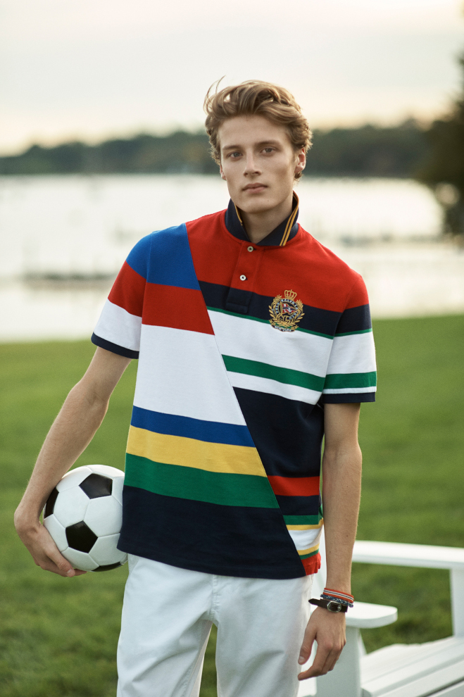 A man is stood in a field holding a football. He is wearing a stiped Ralph Lauren Rugby shirt