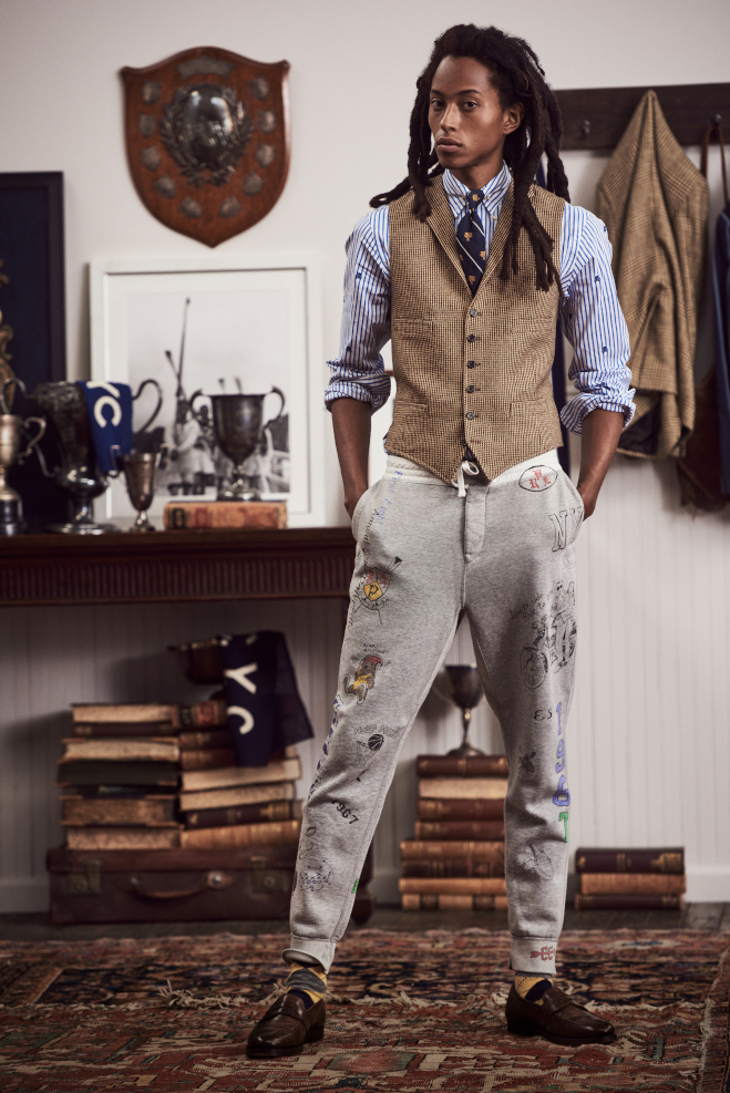 A man is stood in a school room wearing a brown waistcoat, blue striped shirt and light grey jeans. All by Ralph Lauren. 