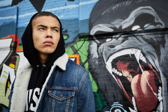 A man looks off to the right wearing a black Diesel hoodie with the hood up and a blue denim jacket. 