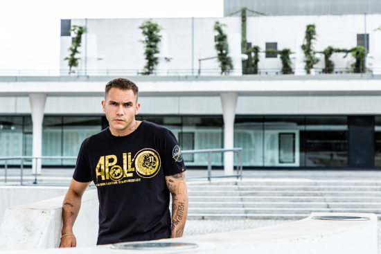 A man is leaning against a fence looking at the camera. He is wearing a black Alpha Industries t-shirt with a gold pattern on the front. 